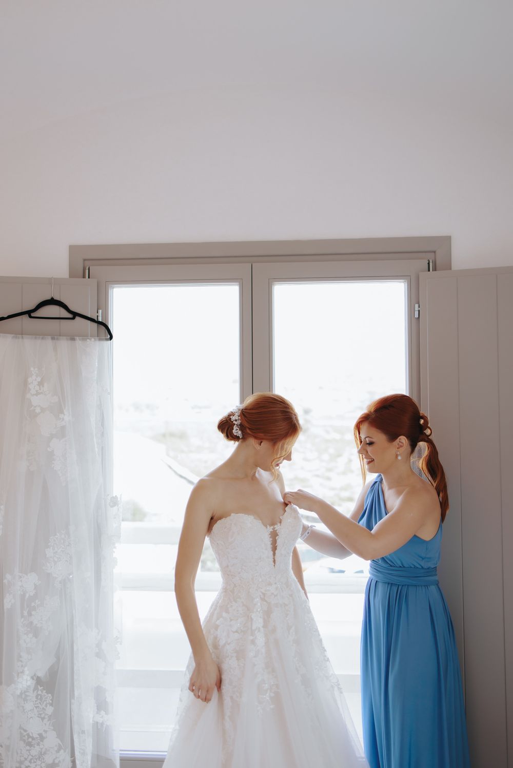 A woman in a blue dress helps adjust a white lace wedding gown by a bright window.