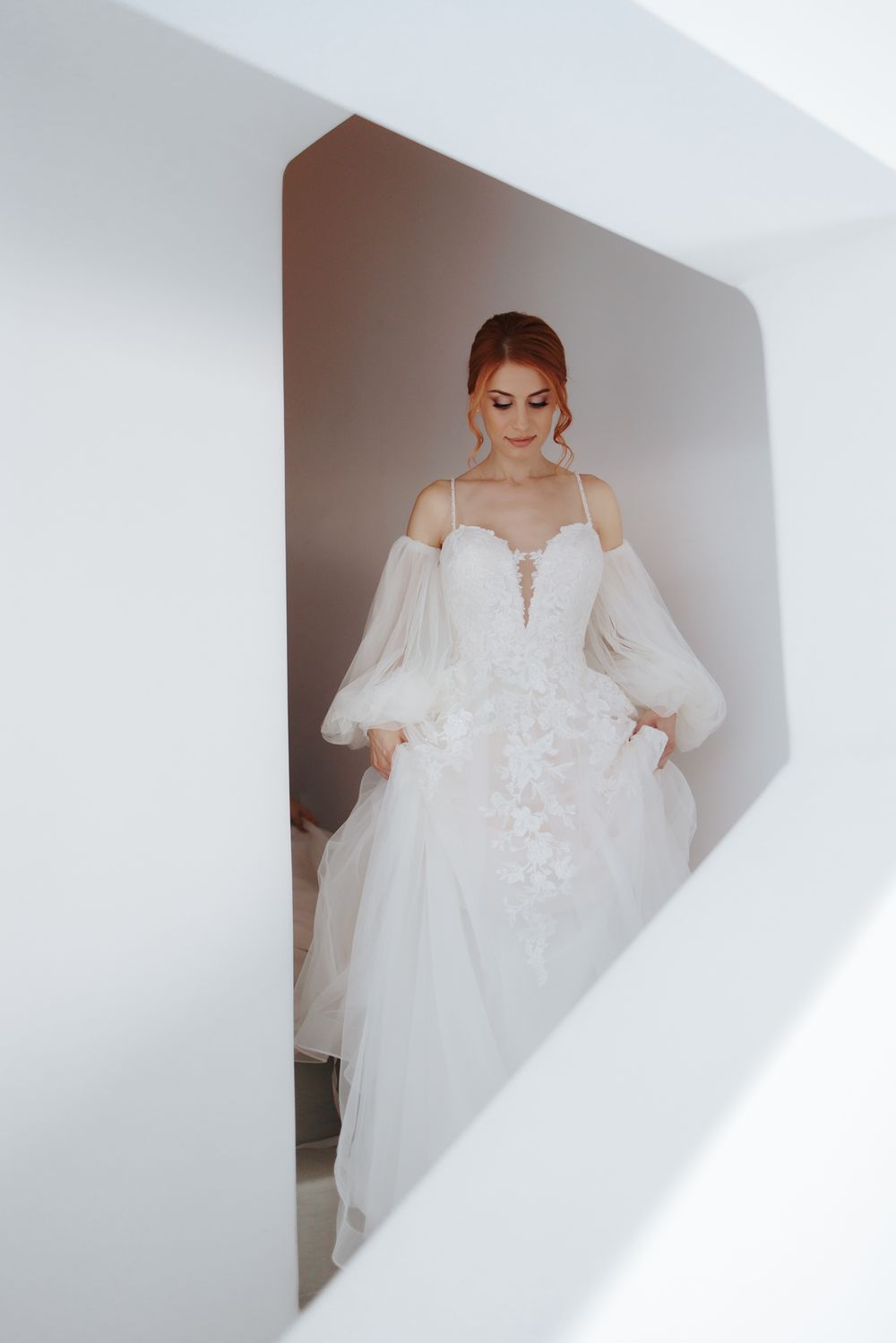 Elegant off-shoulder wedding gown with billowing sleeves, lace details and plunging neckline framed by white walls.
