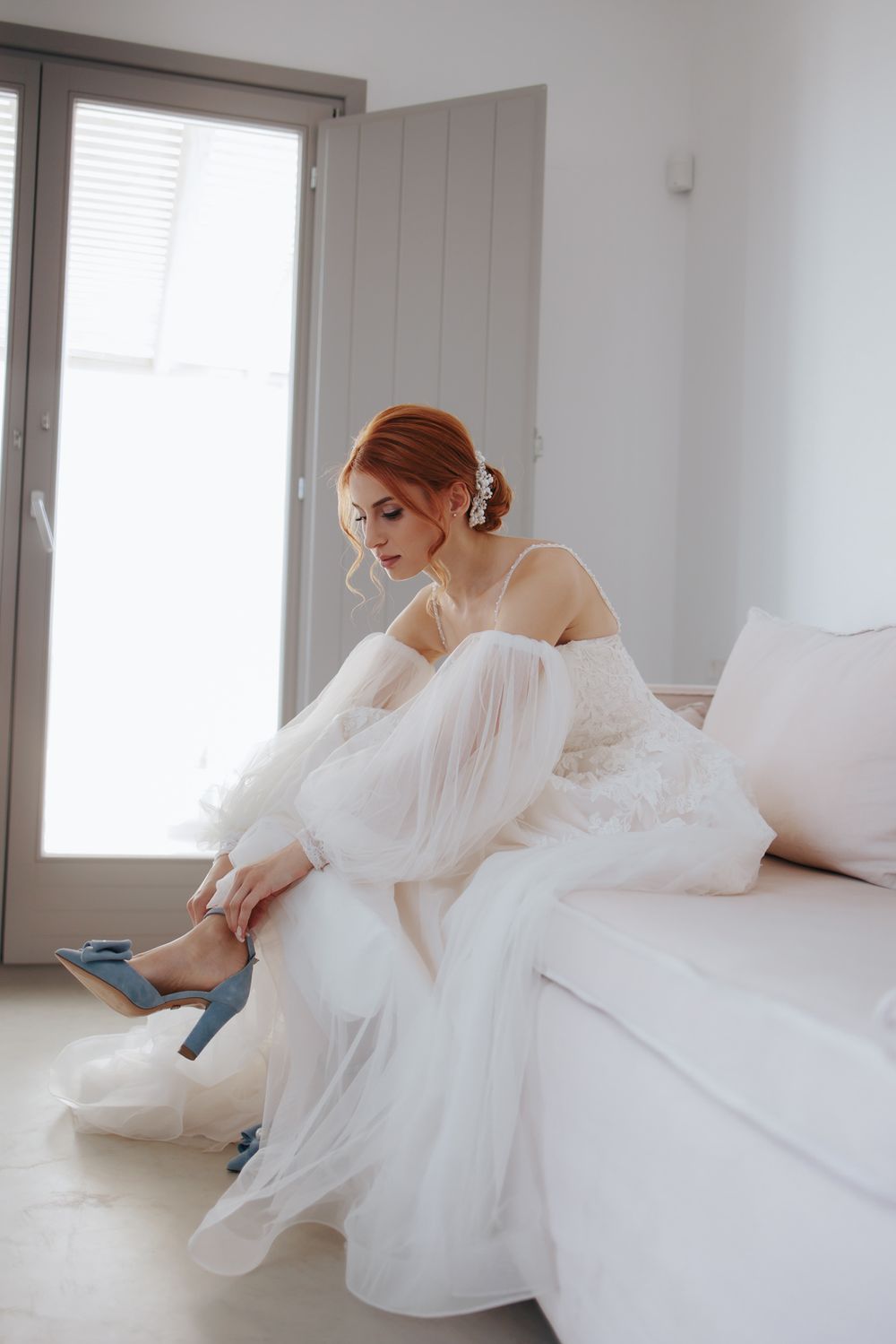 Someone in an ethereal white tulle wedding dress sits on a white sofa while putting on blue heels.