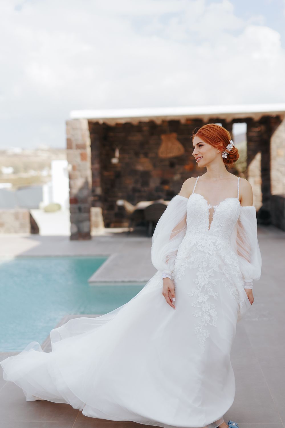 Elegant white wedding dress with off-shoulder puff sleeves and lace details flows gracefully by a poolside terrace.