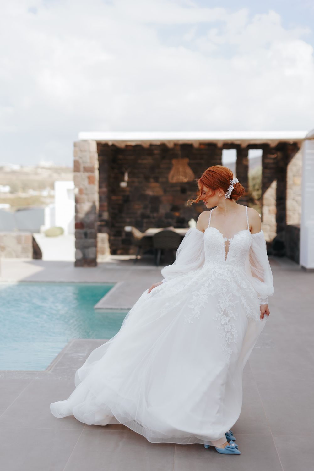A person in a flowing white wedding gown with long sleeves stands by a modern pool deck with blue sneakers.