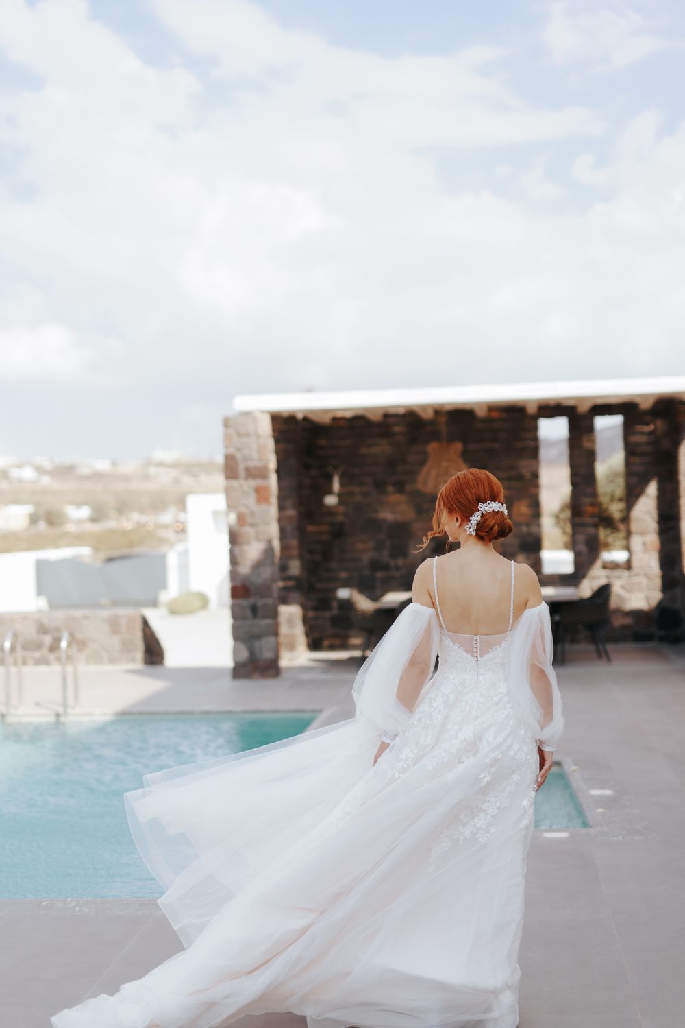 A bride in a flowing white dress with sheer sleeves stands by a pool overlooking a scenic cityscape.