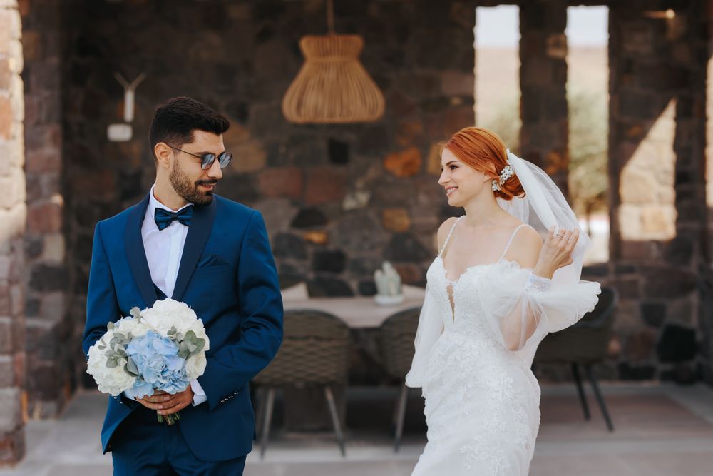A bride in white lace dress and groom in navy suit share a joyful moment while holding blue hydrangea bouquet.