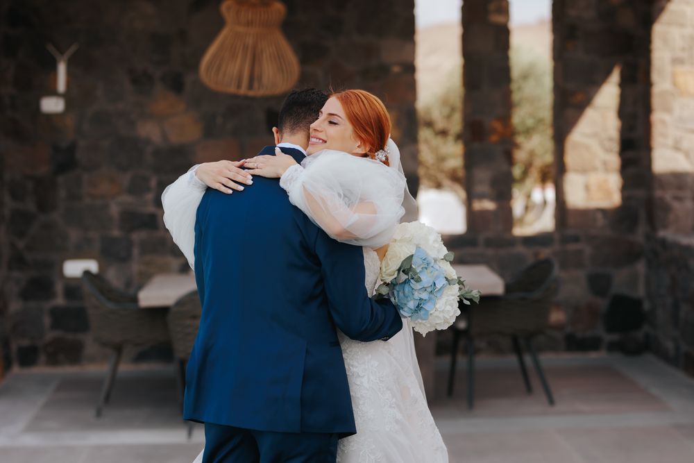 A romantic embrace between newlyweds is captured outdoors with light blue floral bouquet against a rustic stone backdrop.