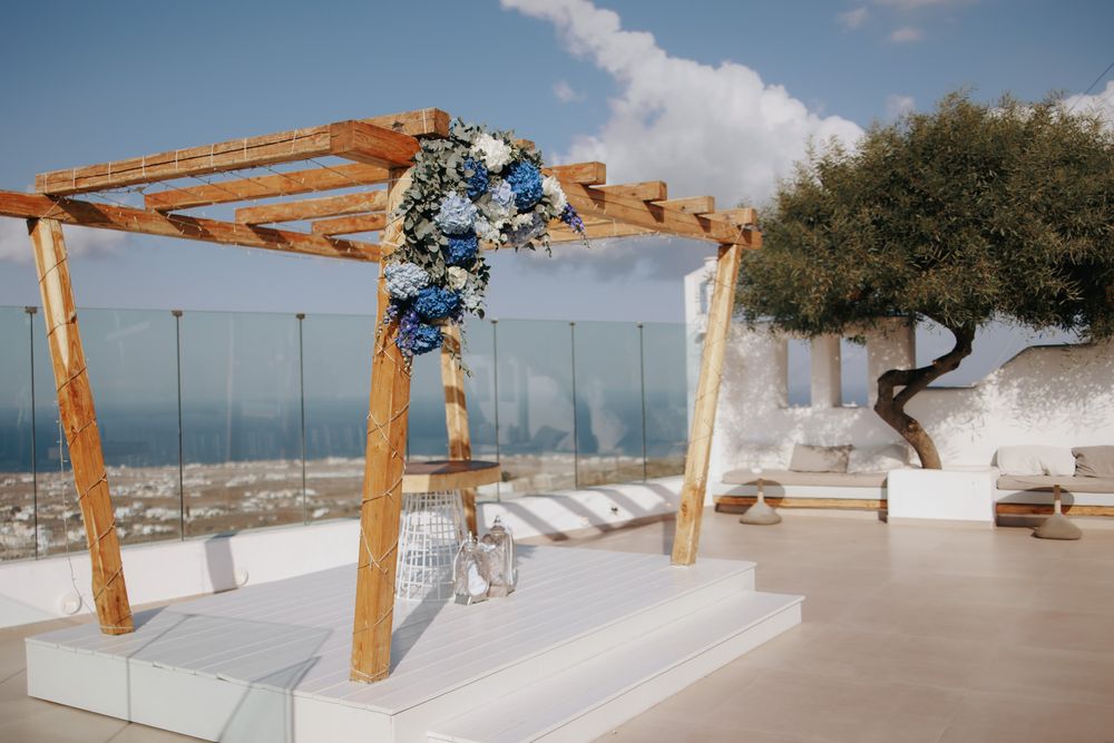A wooden wedding arch decorated with blue flowers stands on a white terrace overlooking the Mediterranean Sea.