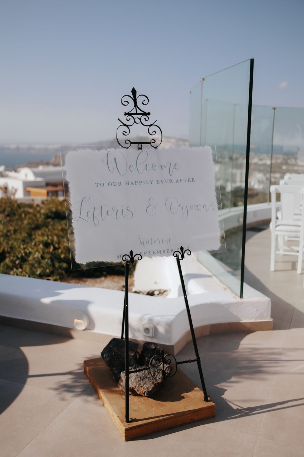 An elegant white wedding welcome sign displayed on a decorative metal stand against a scenic coastal background.