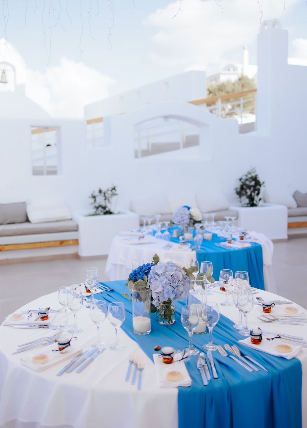 Elegant wedding reception table setting with white linens, bright blue runner and crystal centerpieces on white terrace.