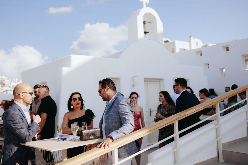 Wedding guests celebrate at an outdoor reception by a traditional white Greek church with bell tower in Santorini.