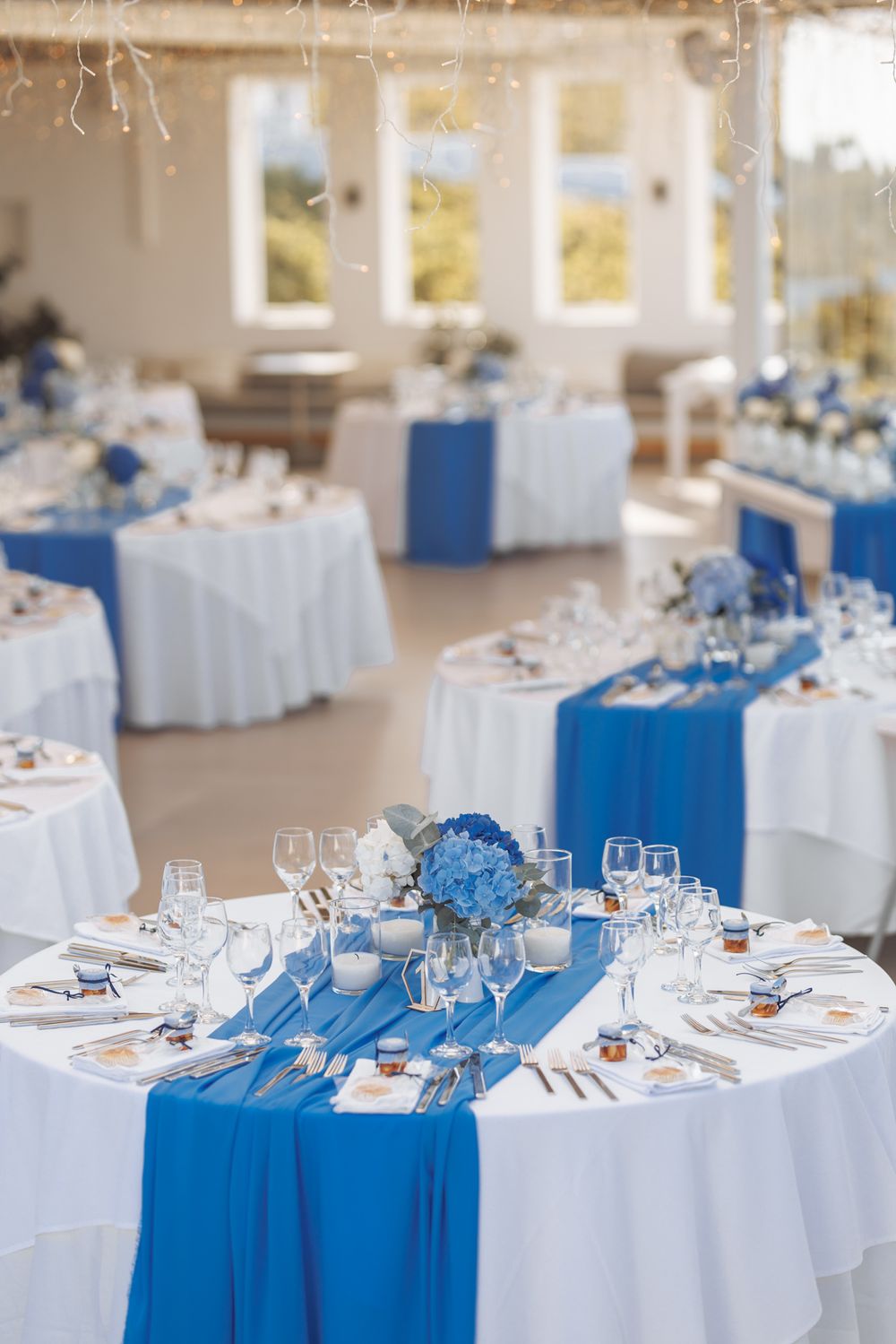 Elegant wedding reception tables decorated with blue runners and hydrangea centerpieces in a bright venue with windows.