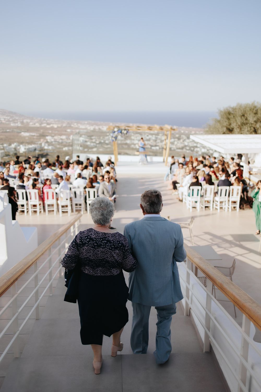 Two people walking down outdoor steps toward a scenic seaside wedding ceremony with guests seated in white chairs.