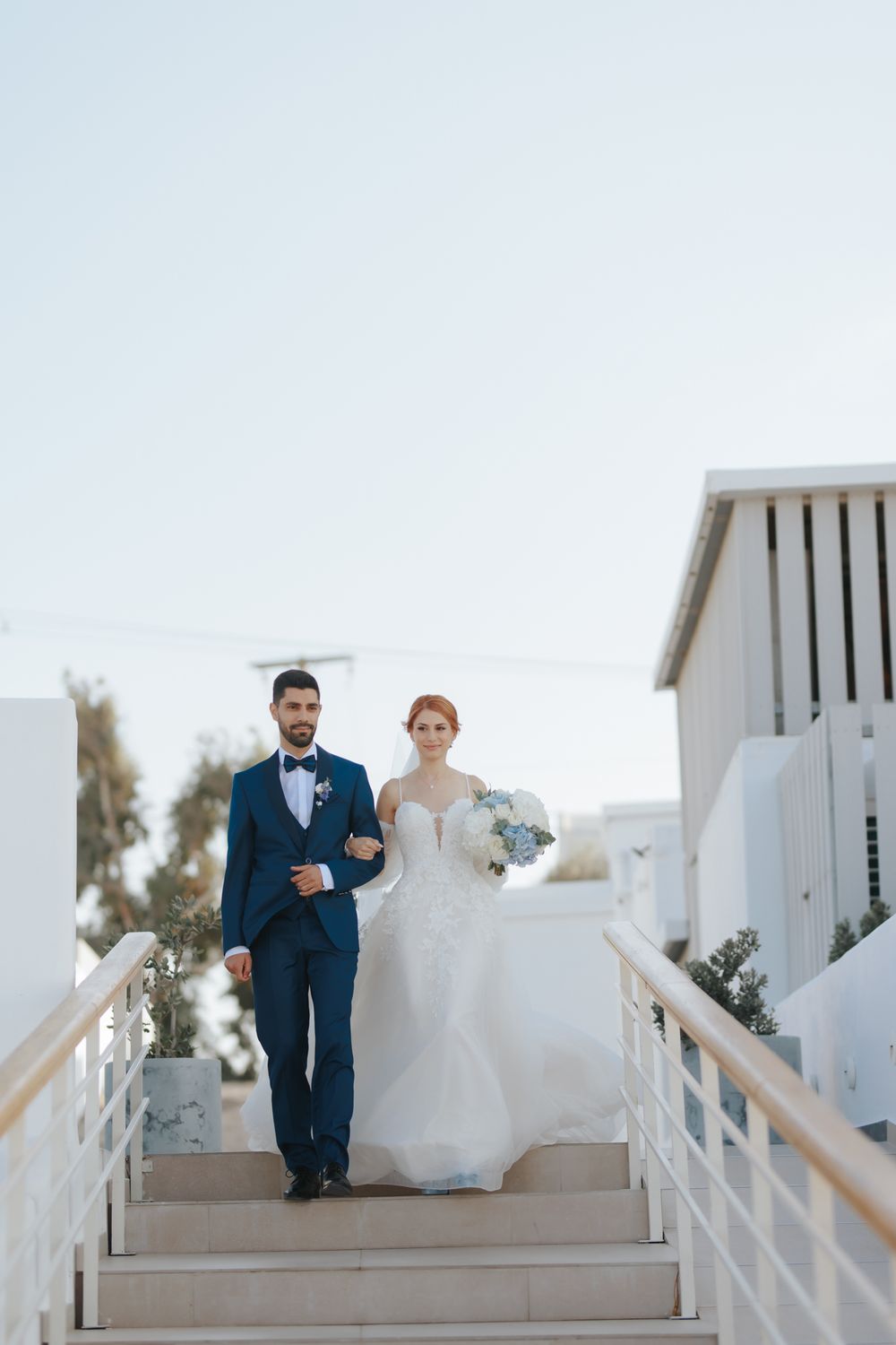 A couple walks down white stairs at their modern coastal wedding venue, wearing navy and white formal attire.