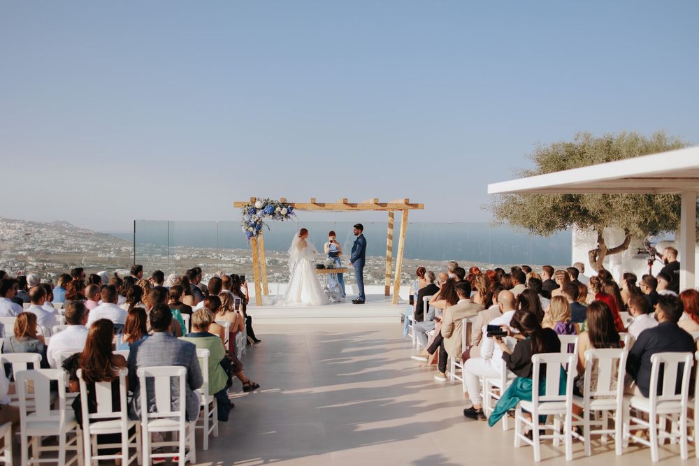 A beautiful outdoor wedding ceremony overlooking the sea with guests seated in white chairs and a wooden arch.