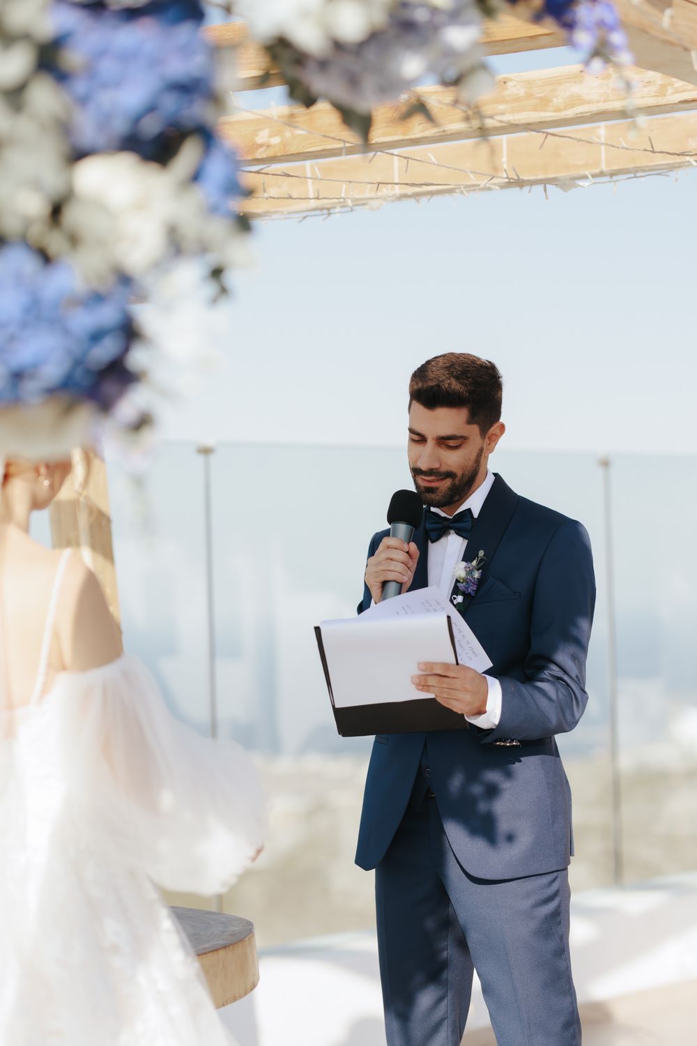 A groom reads from a paper during an outdoor wedding ceremony decorated with blue and white floral arrangements.