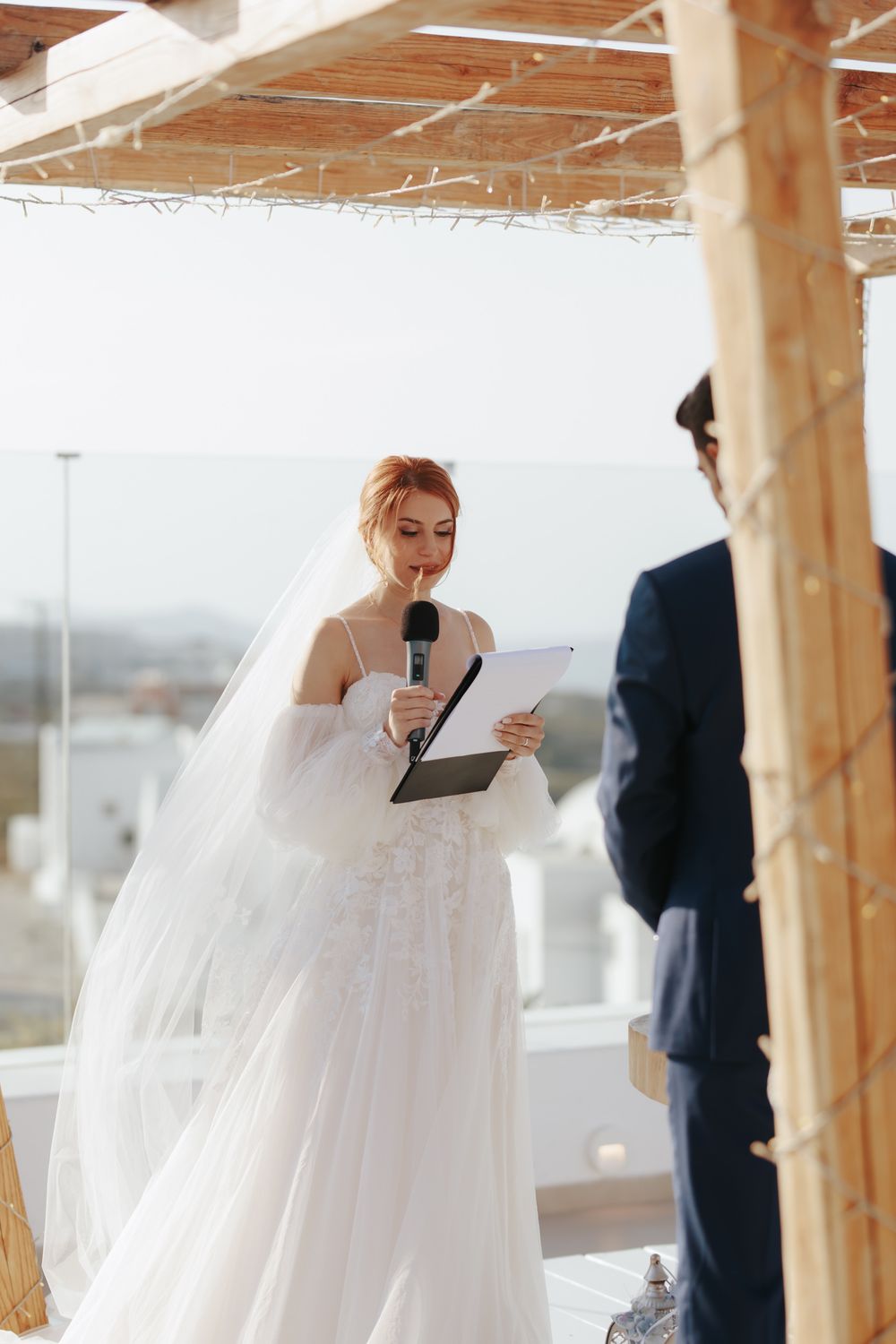 A bride in a flowing white wedding dress reads from a document during an outdoor ceremony.