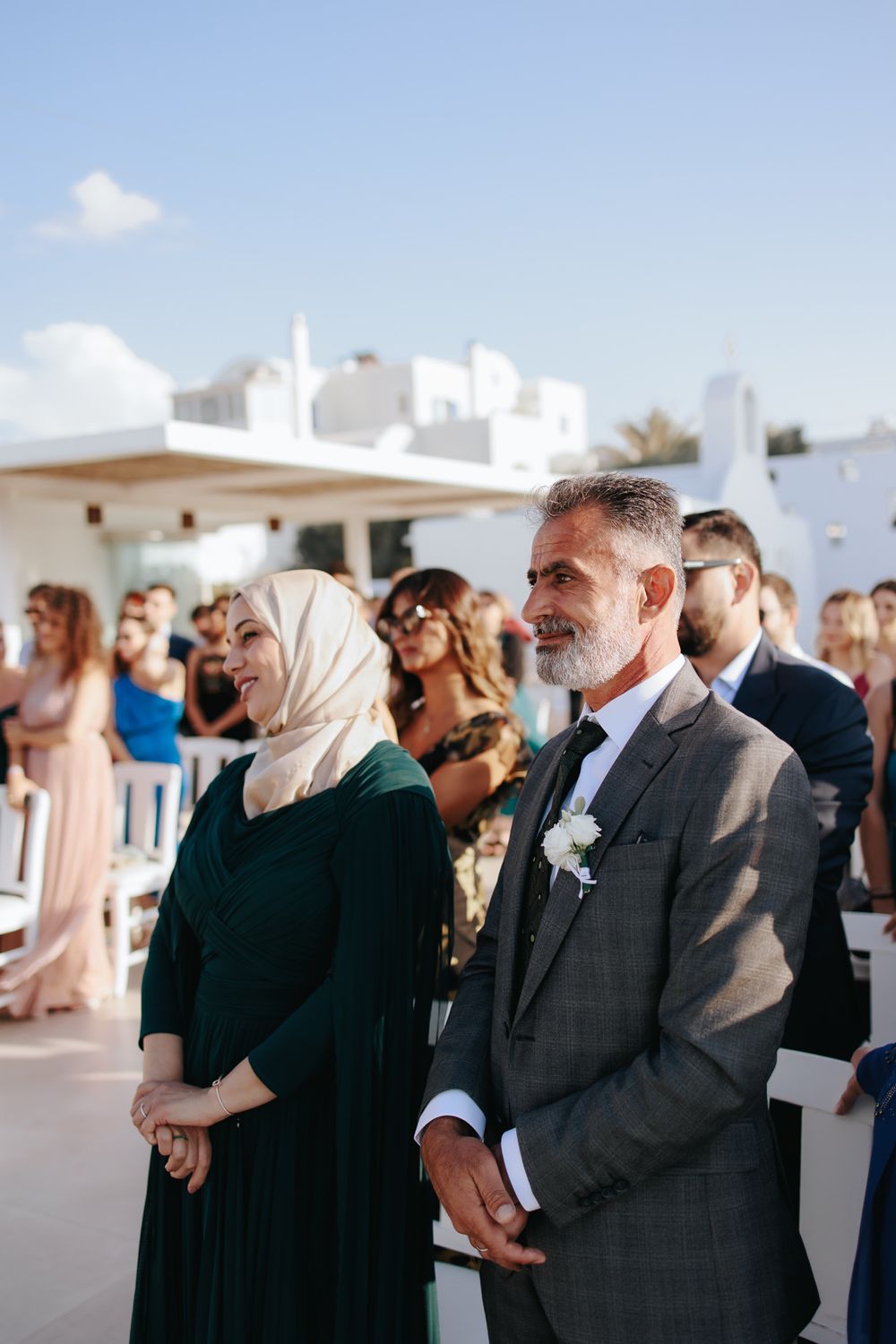 Wedding guests watch ceremony outdoors under blue sky at elegant venue with white buildings in background.