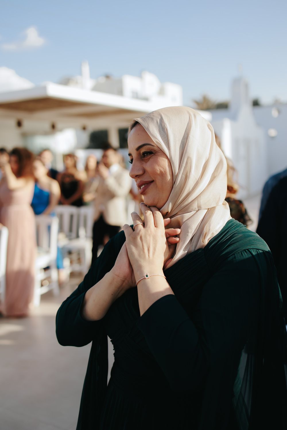 A guest in a cream hijab and dark green dress stands joyfully at an elegant event venue.