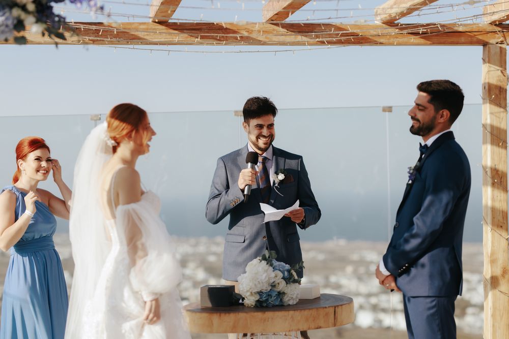 Intimate outdoor wedding ceremony with officiant and couple under wooden pergola overlooking coastal mountains.