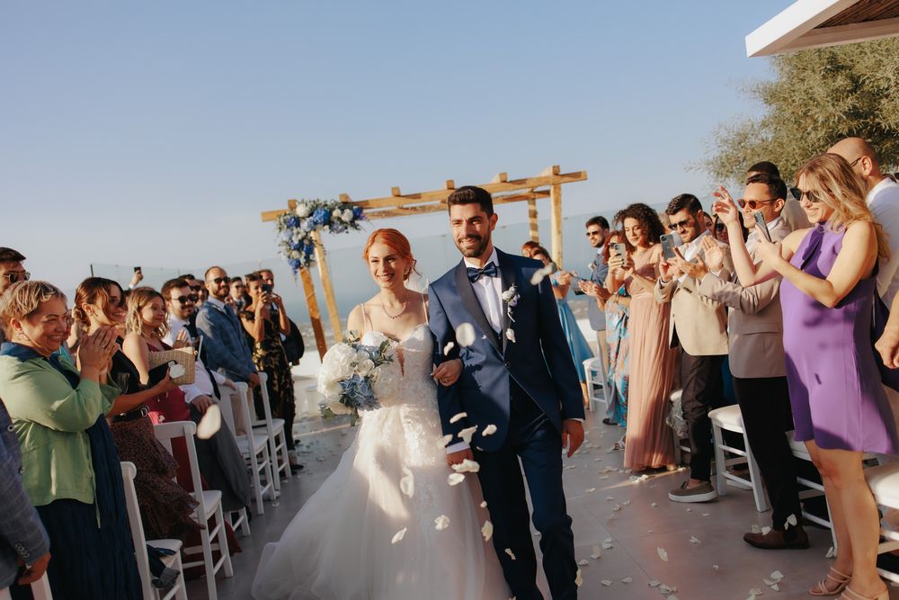A newlywed couple walks down an outdoor aisle while guests take photos at a scenic mountain wedding ceremony.