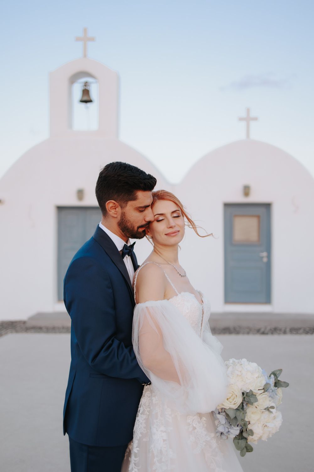 A romantic wedding couple embraces in front of a white Mediterranean church with blue doors and cross details.