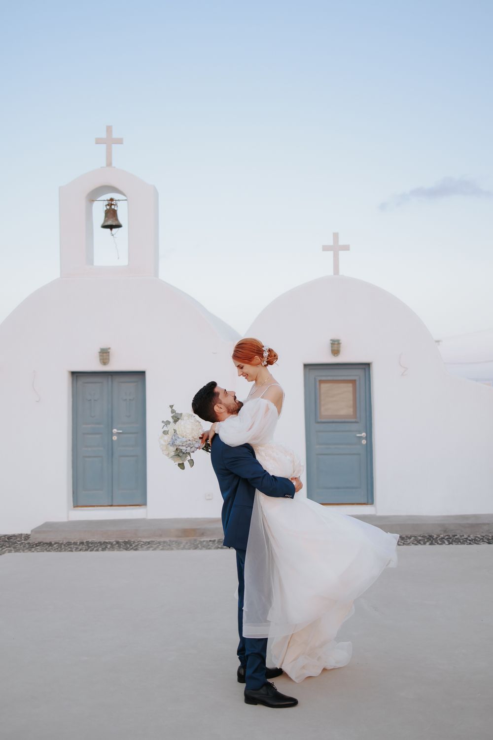 A romantic wedding couple embraces in front of a white Mediterranean church with blue doors and bell tower.