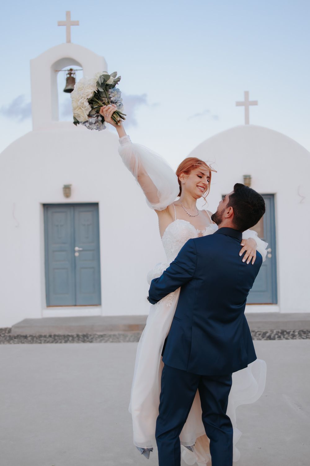 Newlyweds share a joyful dance pose outside a traditional white Greek church with blue doors.