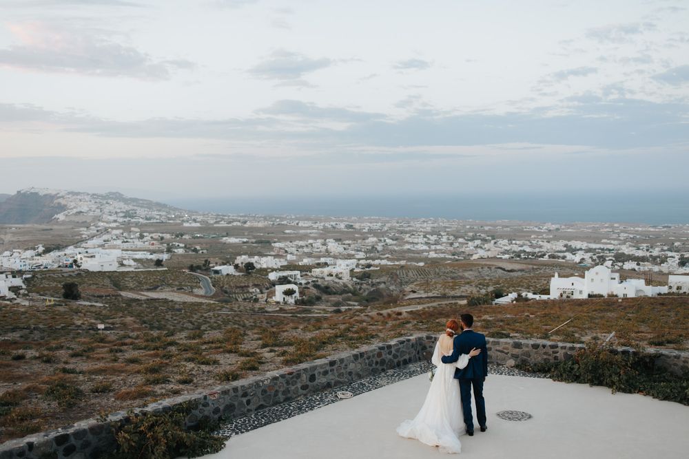 A couple embraces on a stone path overlooking the white buildings and coastal landscape of Santorini, Greece.