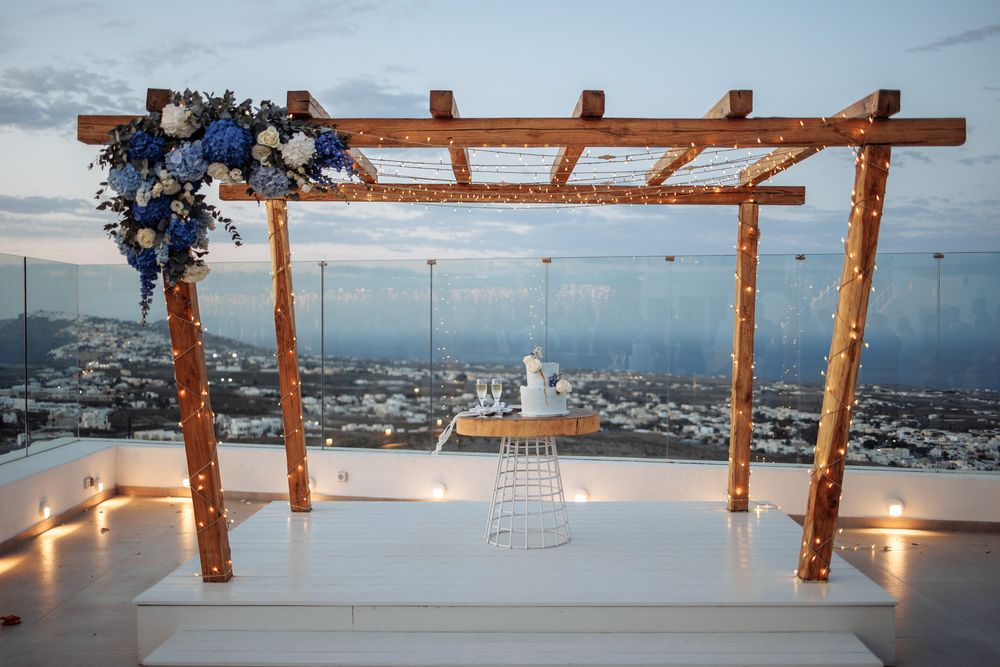 Wooden wedding arch decorated with blue flowers overlooks a coastal city view at sunset with romantic lighting.