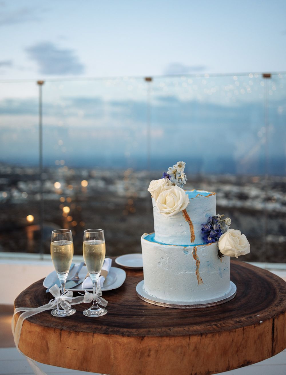 Elegant two-tier white and blue wedding cake with roses on wooden platter alongside champagne glasses at sunset overlooking city.