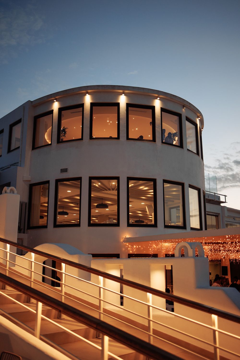 Modern circular building with illuminated windows and ramp entrance during dusk.