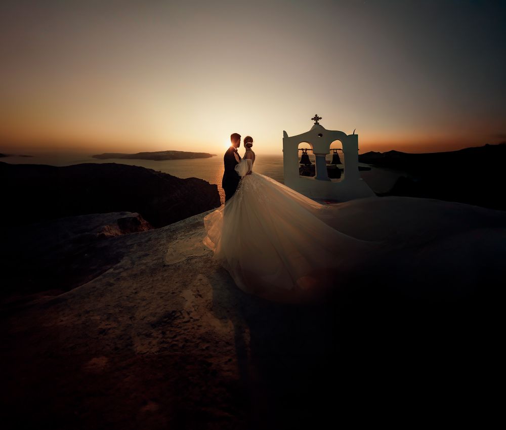 A silhouetted couple stands near a whitewashed church bell tower at sunset on a Greek island.