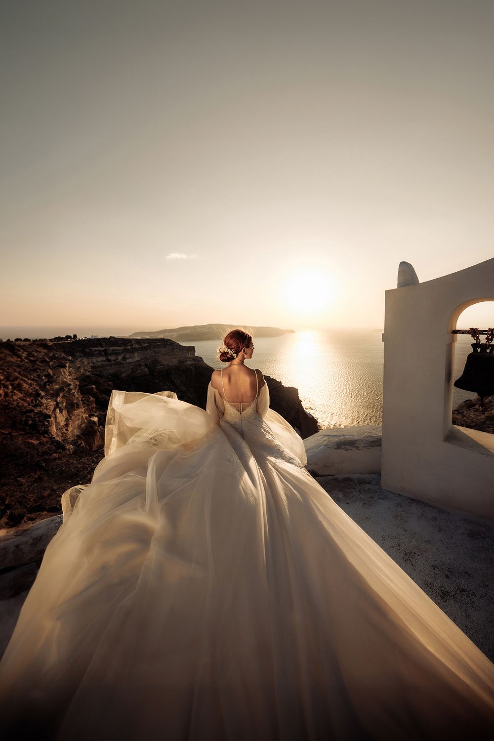 A stunning white wedding dress flows dramatically against a sunset backdrop over the Mediterranean Sea in Santorini, Greece.