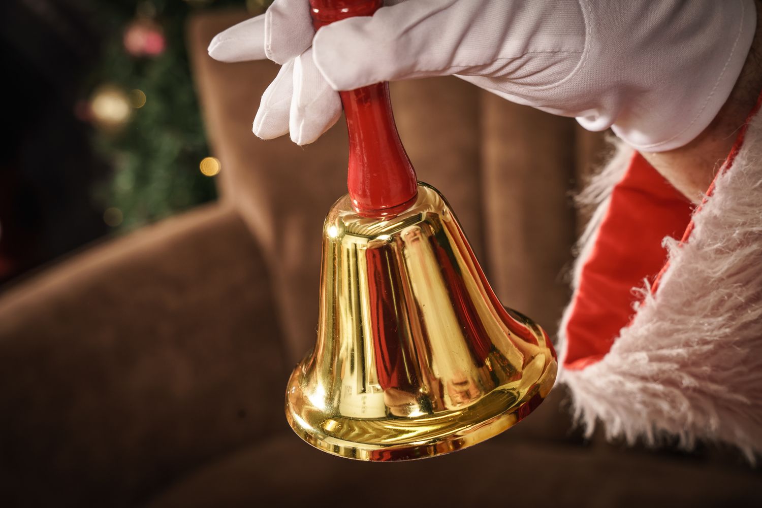 White gloved hand holding a shiny golden Christmas bell against a dark background.