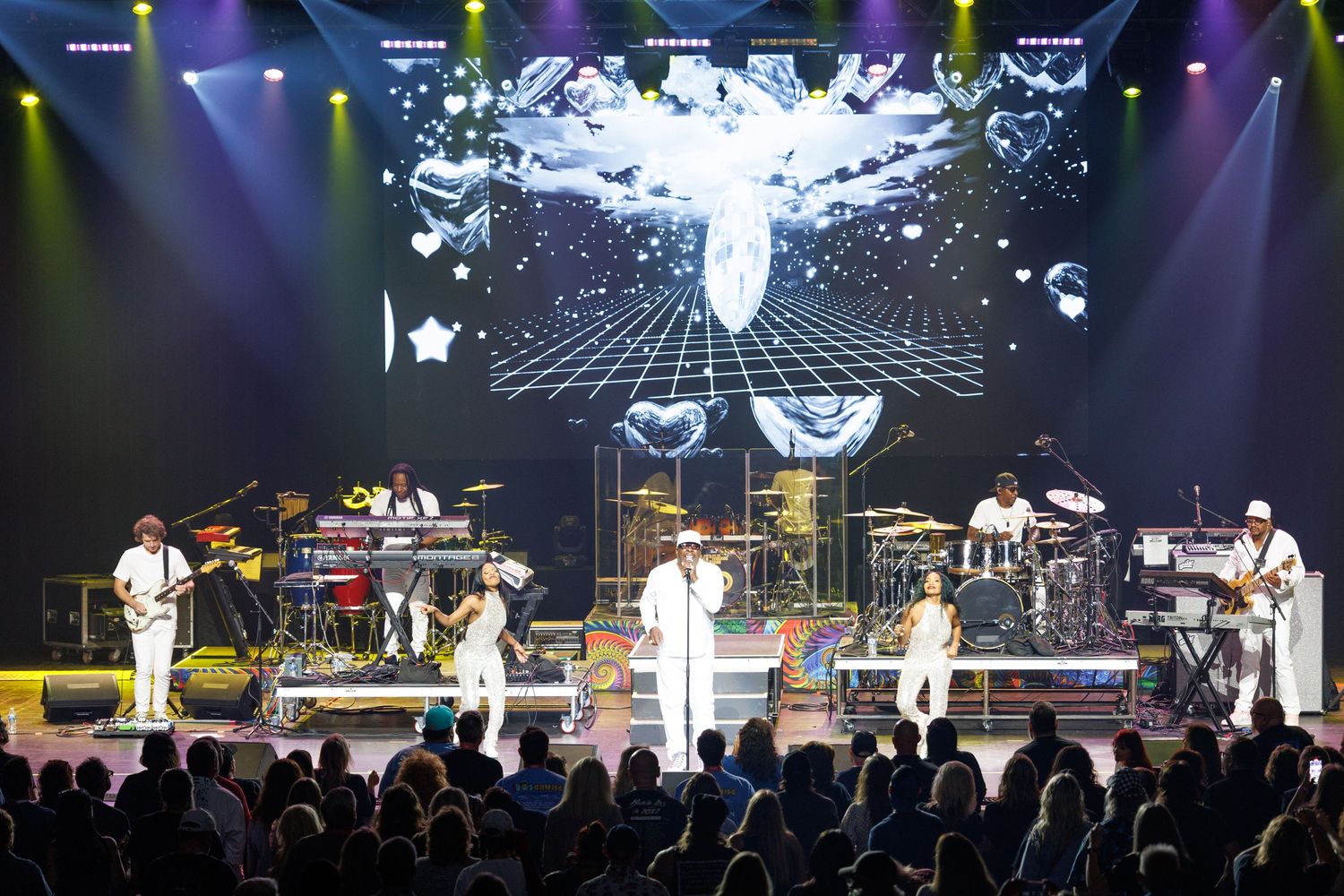 A live concert performance with musicians on stage in white outfits against a dramatic backdrop with projected visuals.