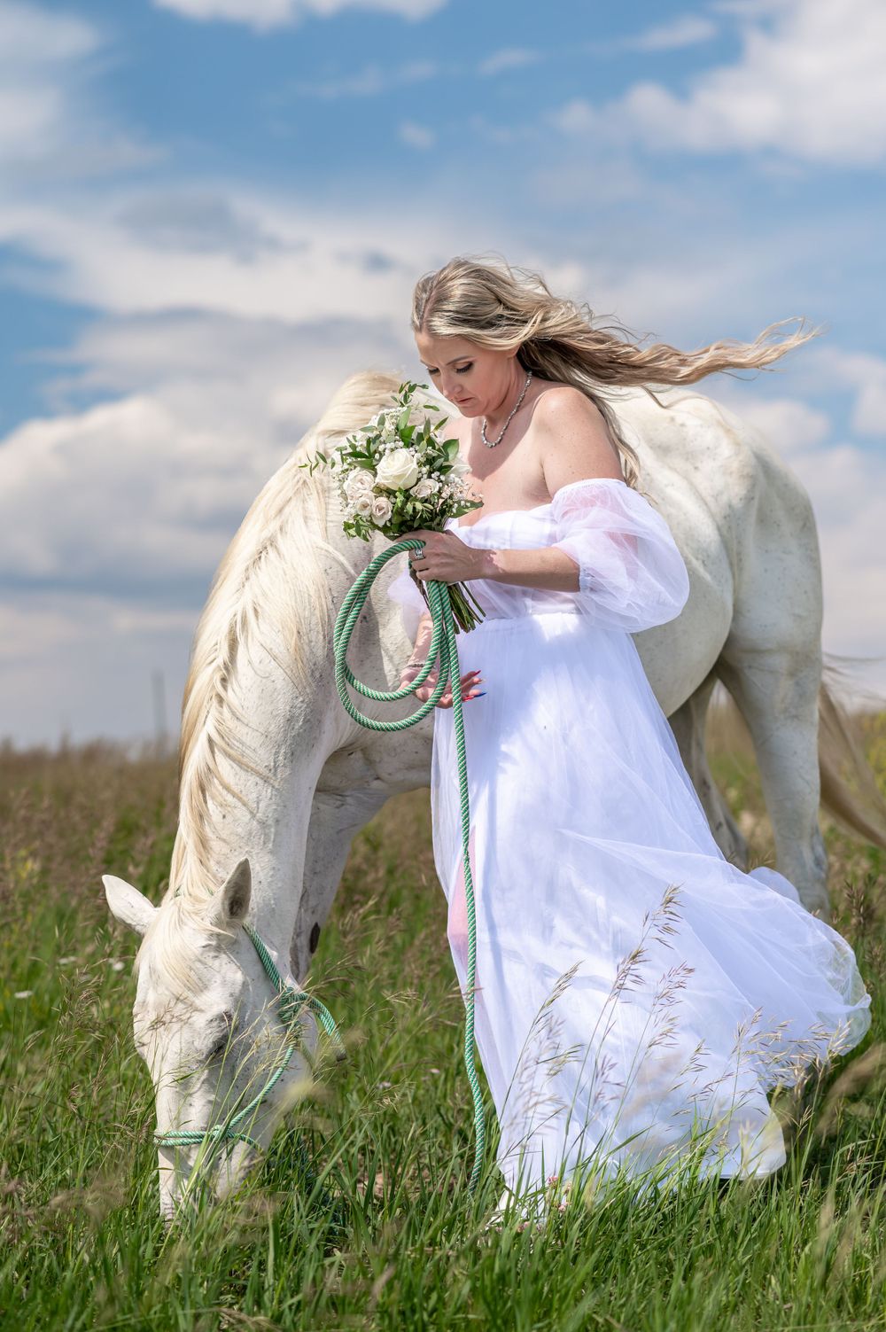 A white horse and figure in flowing white dress stand in a grassy meadow with a bouquet against a blue cloudy sky.