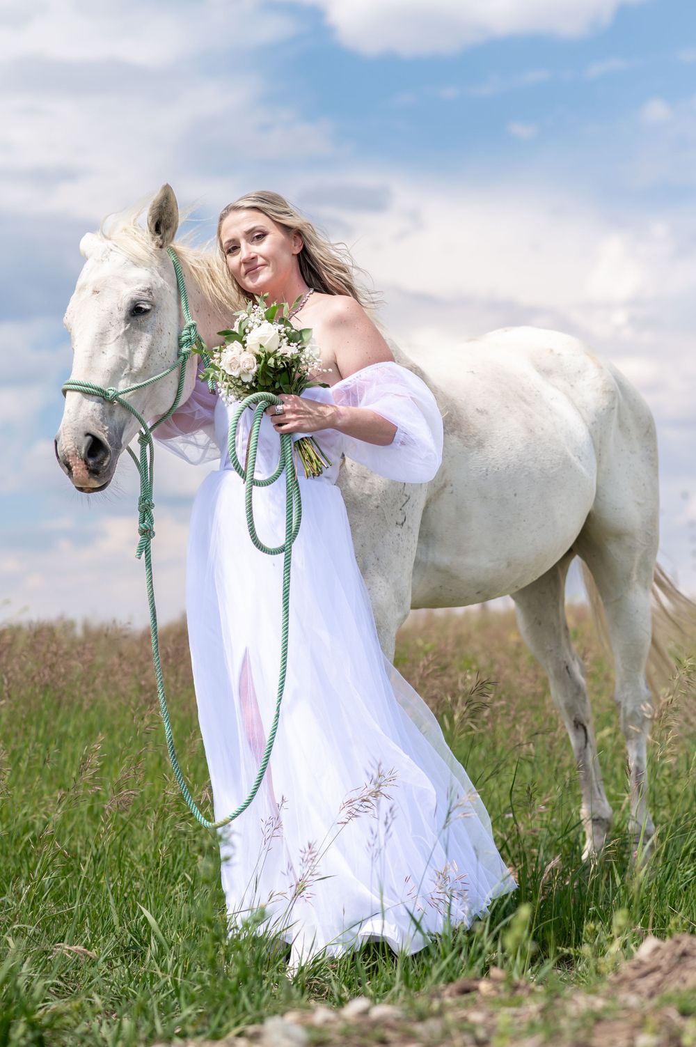 A white horse stands beside someone in a flowing white wedding dress holding flowers in a grassy meadow under blue sky.
