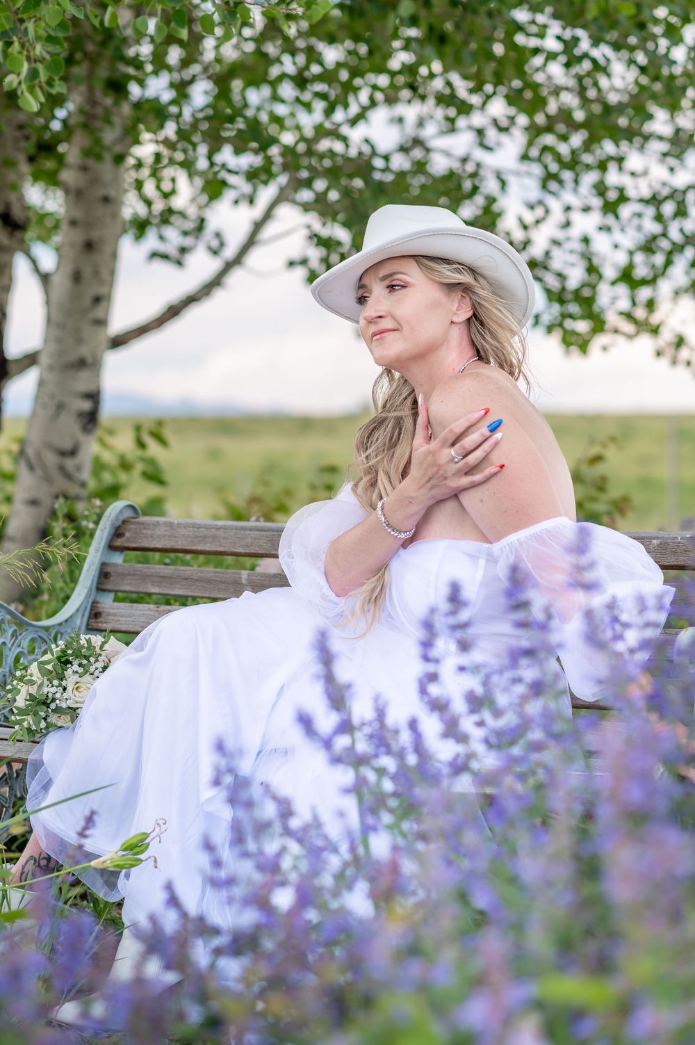 Person in white dress and cowboy hat sits dreamily on bench amid blooming lavender flowers in garden setting.