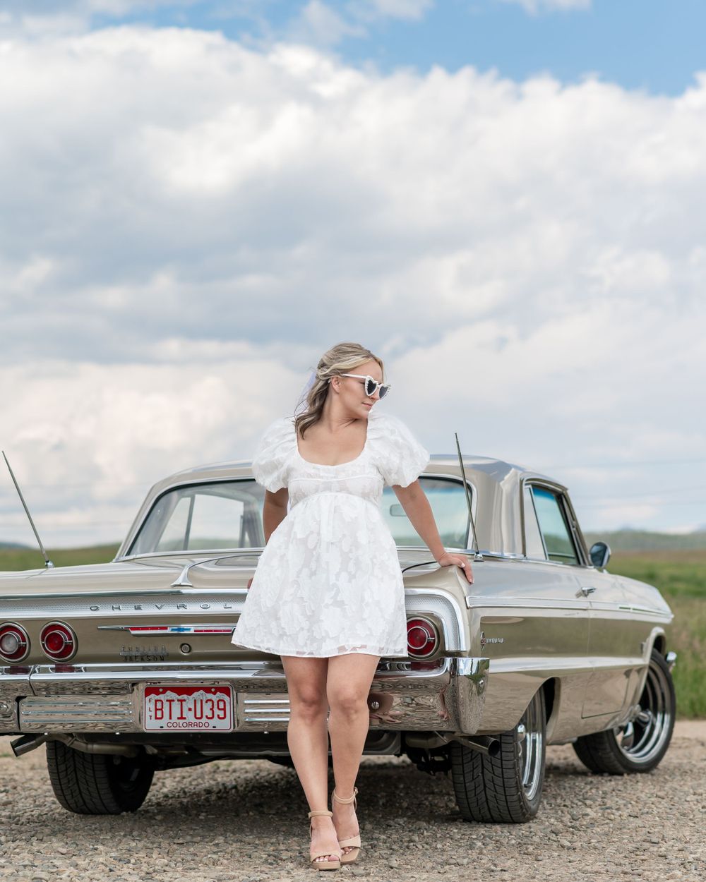 A white dress wearing woman stands beside a vintage 1960s Chevy Impala on a gravel road under cloudy skies.