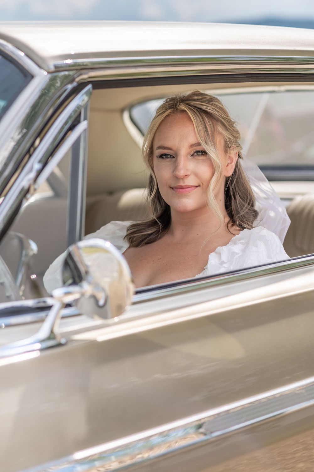 Beautiful bride in white veil smiles from window of classic vintage car on wedding day.