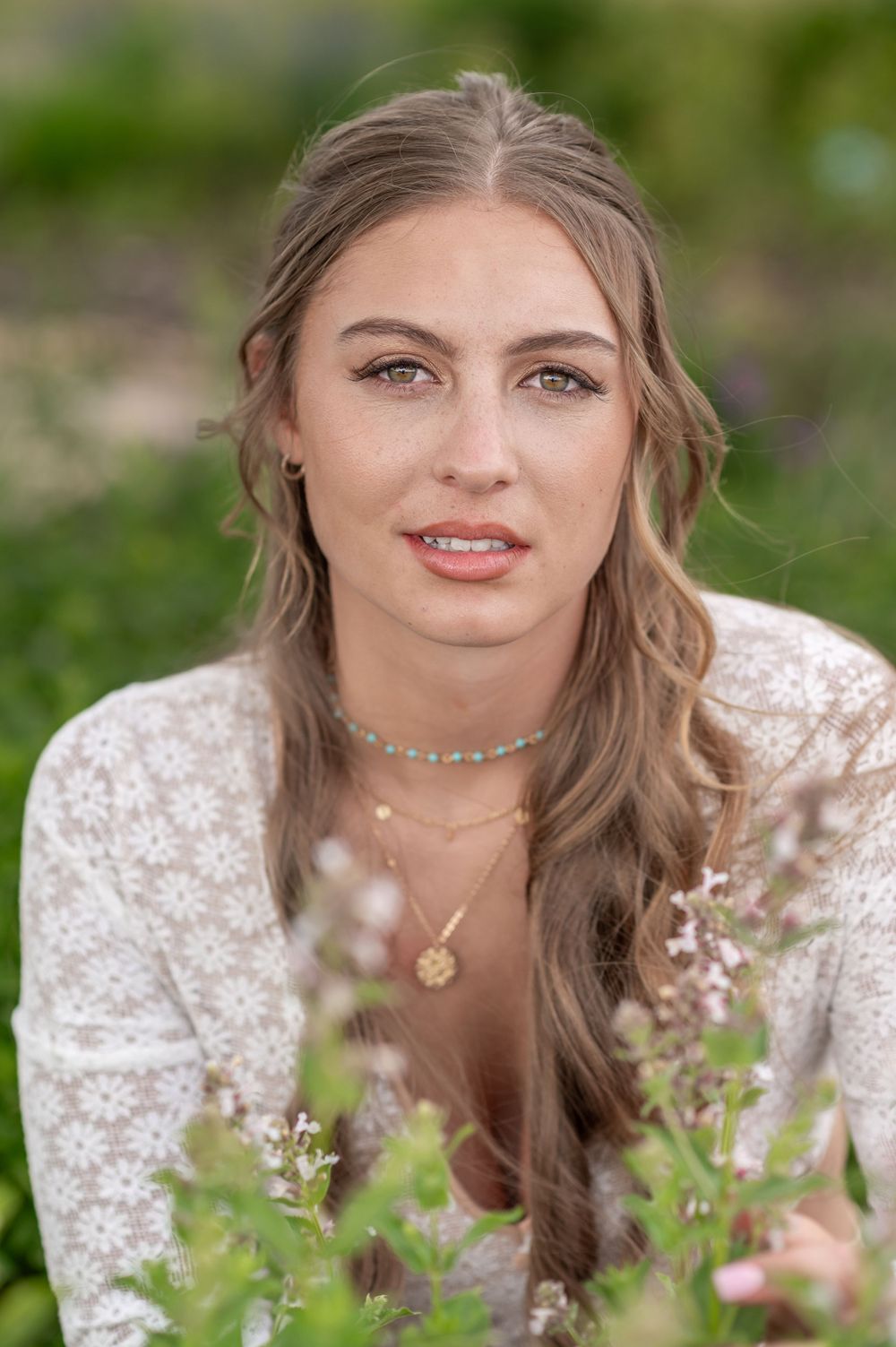 Portrait of someone in a white lace dress sitting among wildflowers wearing layered necklaces with soft blurred background.