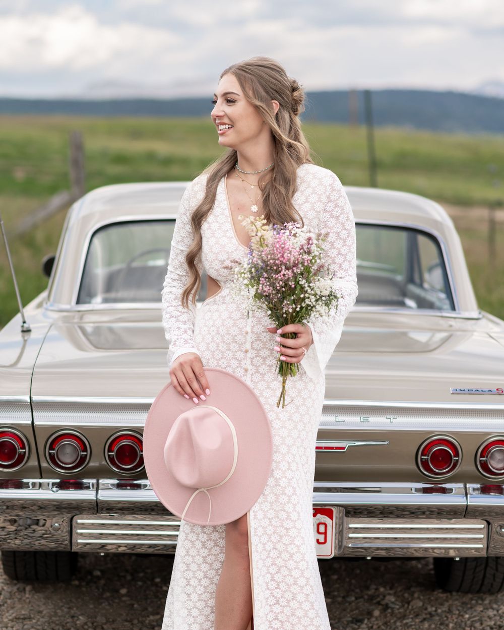 Person in vintage-style lace wedding dress holding wildflower bouquet poses with pink hat by classic Chevrolet Impala.