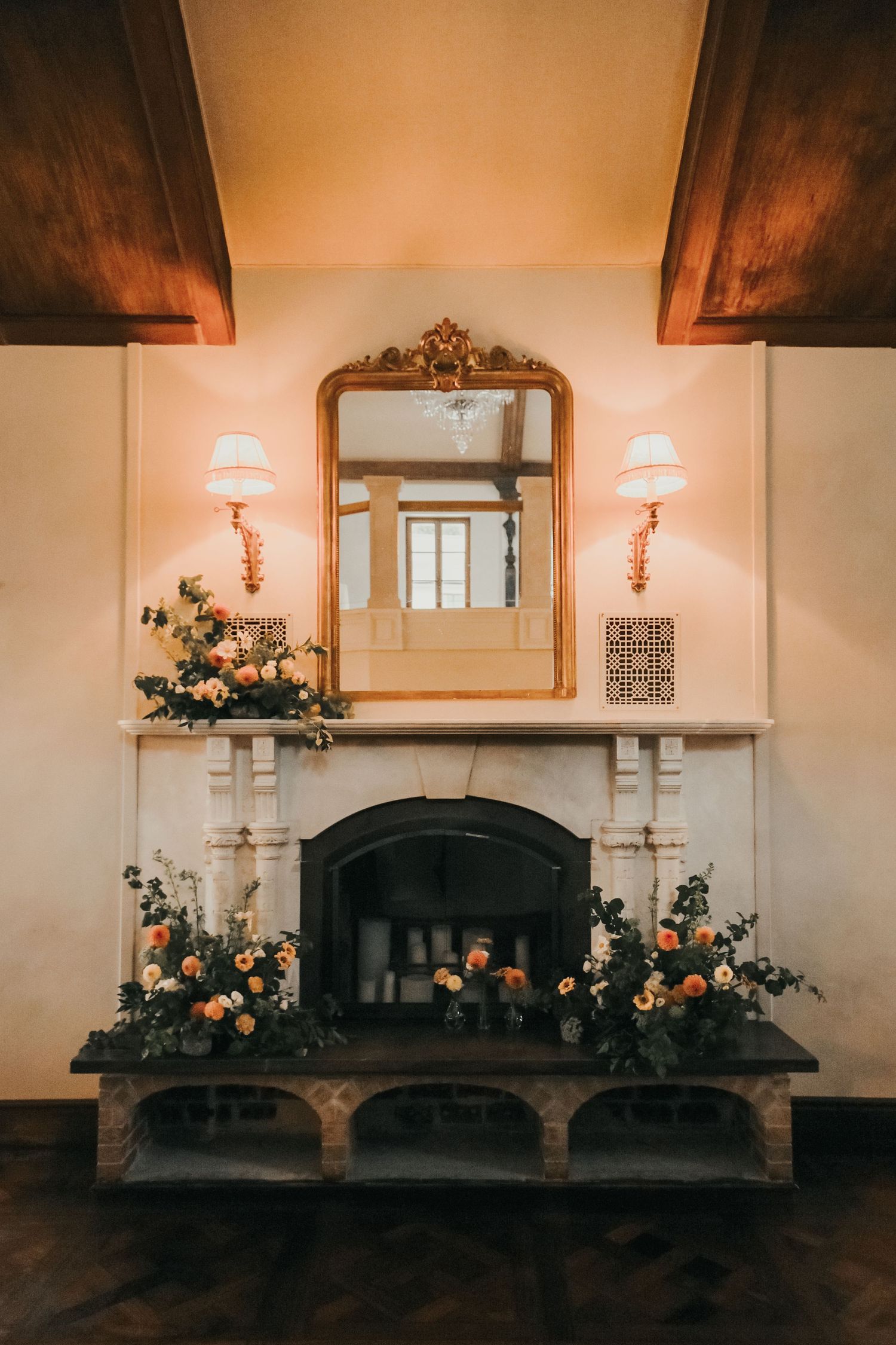 A decorative mantel with candles, floral arrangements, and a mirror in warm lighting creates an elegant wedding reception setting.