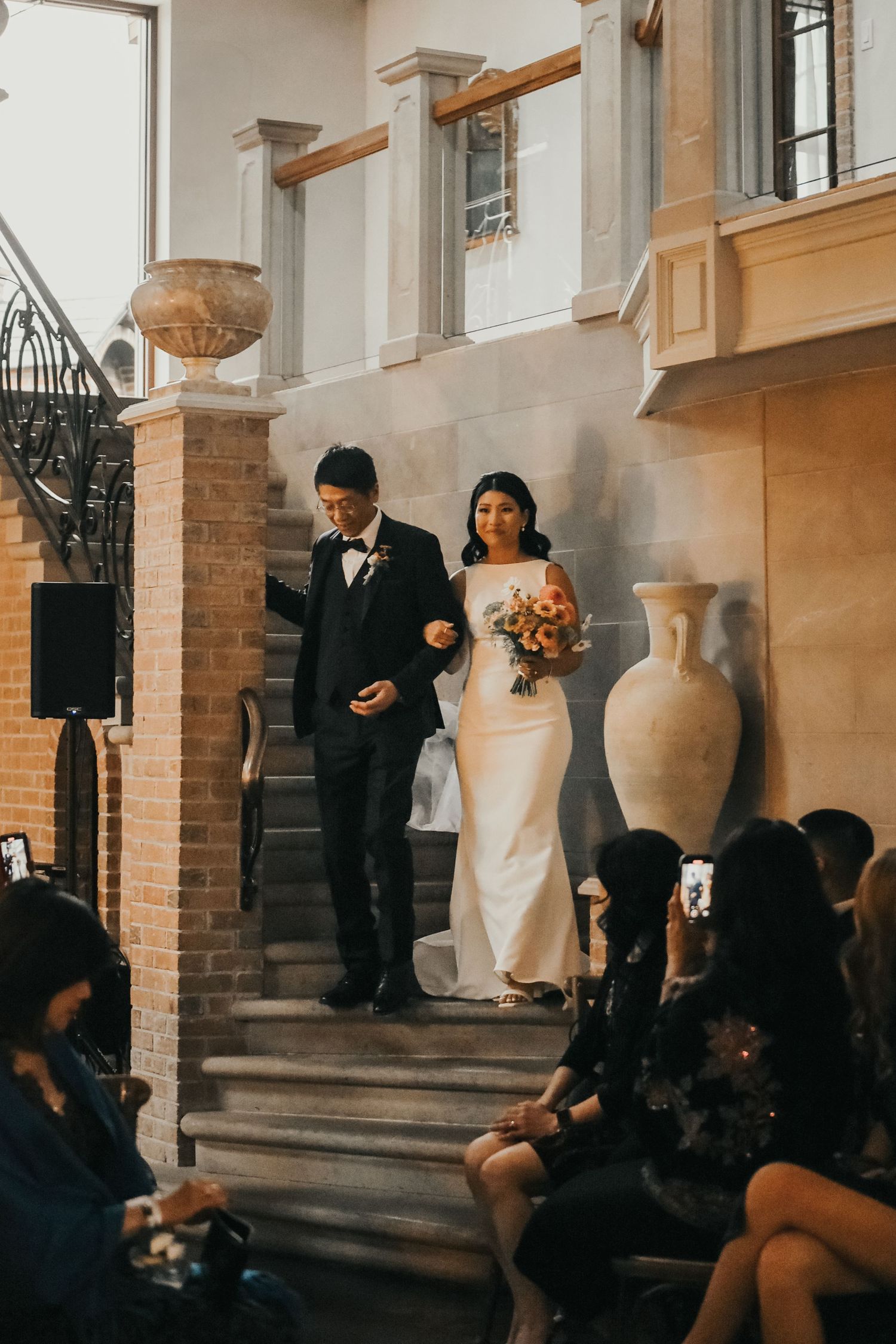 Wedding ceremony on stone steps at an elegant venue with decorative urns and brick architecture.