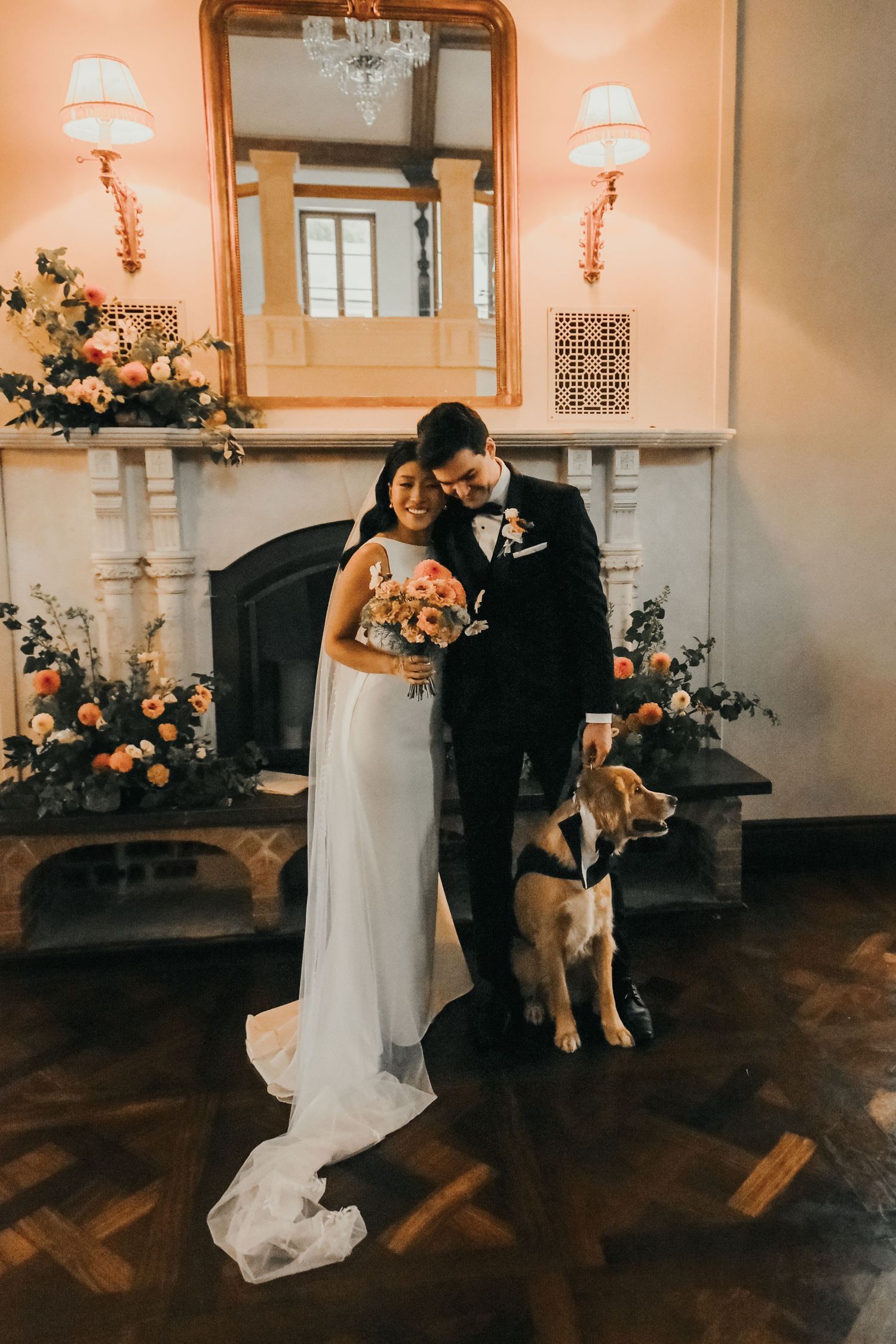 An elegant wedding couple stands by a fireplace in warm lighting with their brown dog and floral decorations.