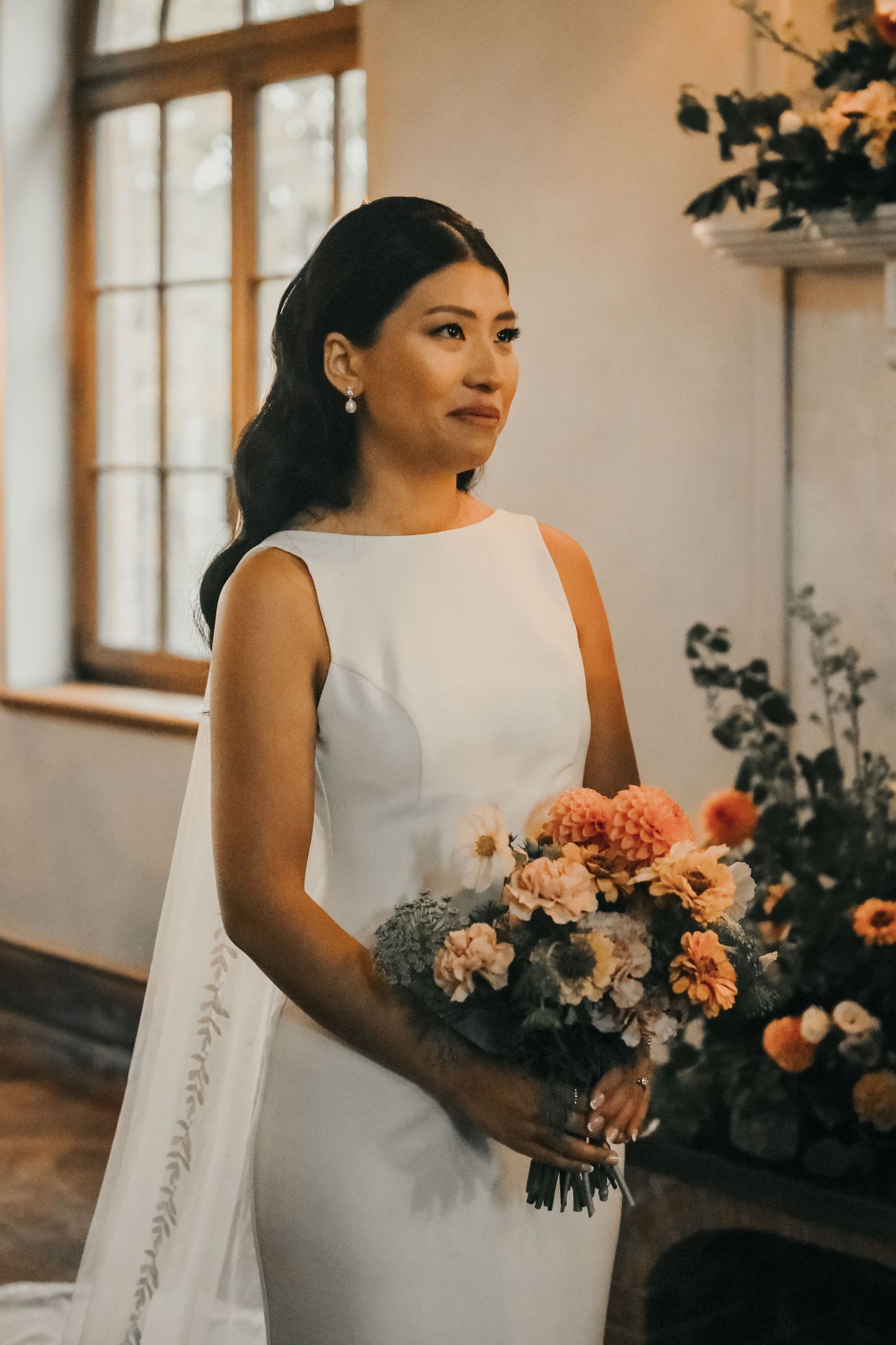 Classic white bridal gown with flowing veil and peach floral bouquet shown in elegant wedding venue setting.