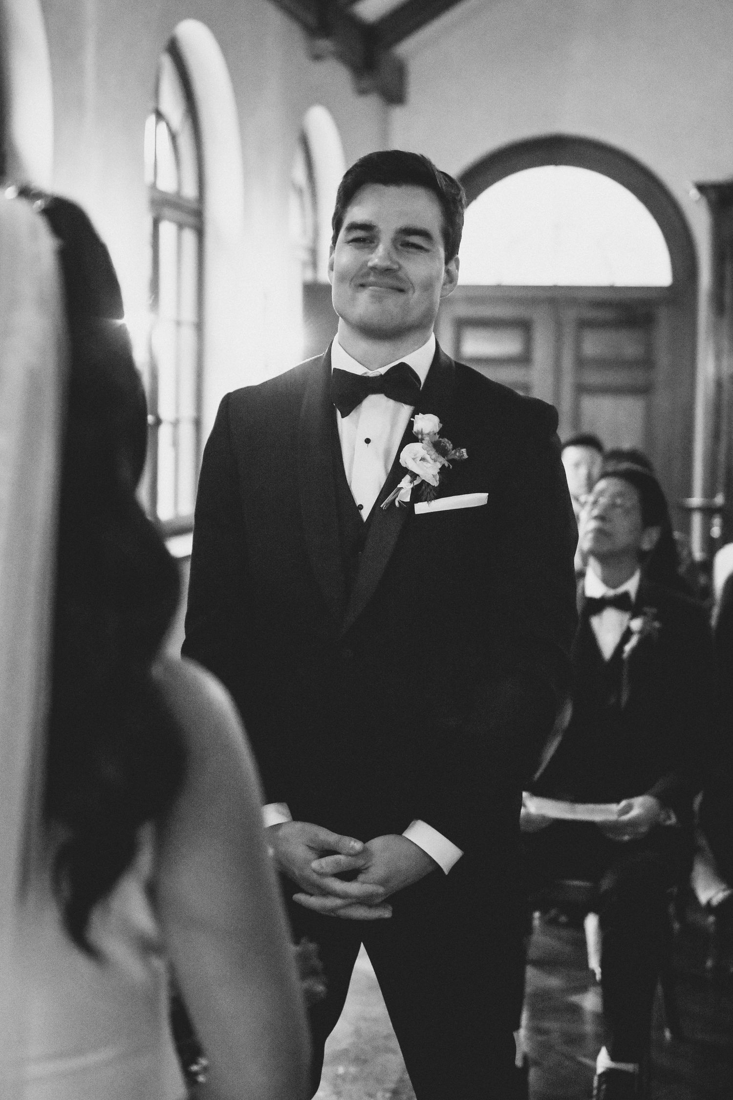 A groom smiles adoringly at his bride during their indoor wedding ceremony, captured in elegant black and white.