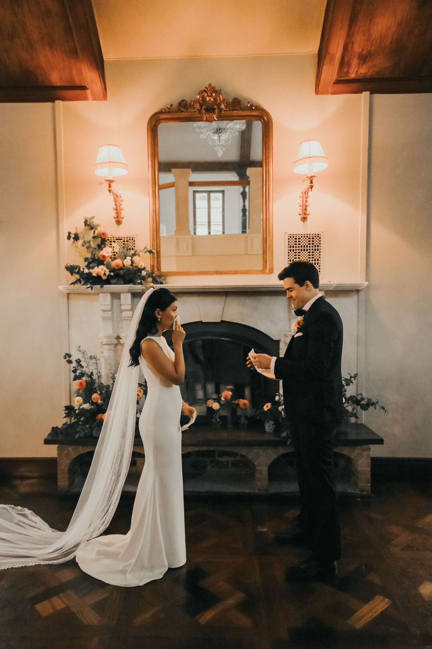 Newlywed couple share their first dance in a warmly lit indoor venue with wall sconces and hardwood floors.