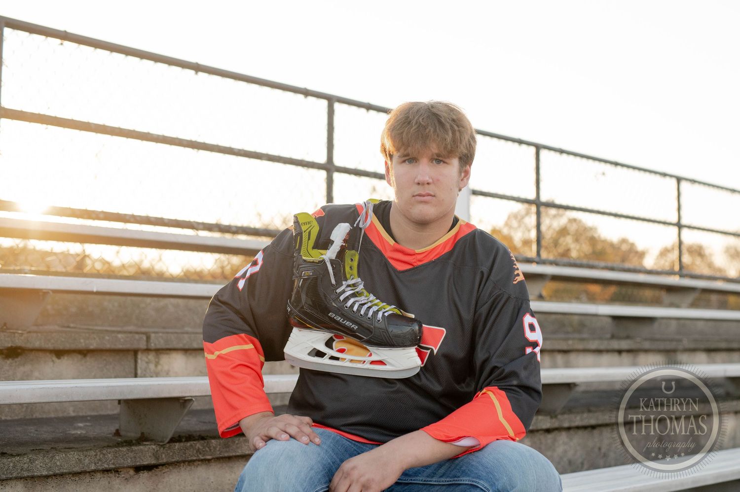Athlete sits on bleachers at sunset wearing a black and orange sports jersey with ice skates.