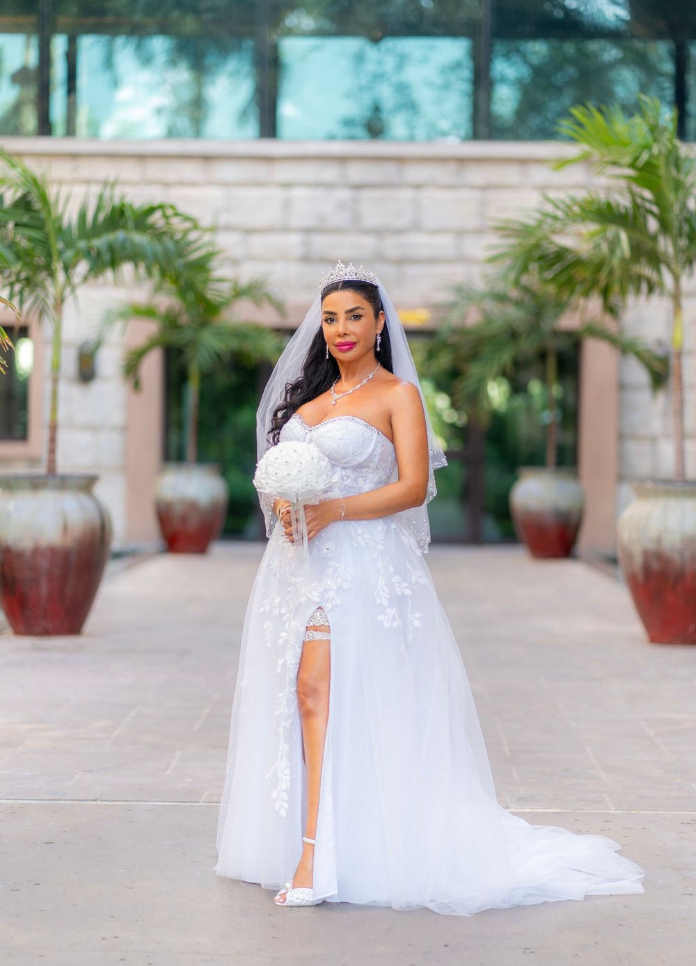 A bride poses outdoors in a beautiful strapless white lace wedding gown with side slit and flowing train near tropical palms.