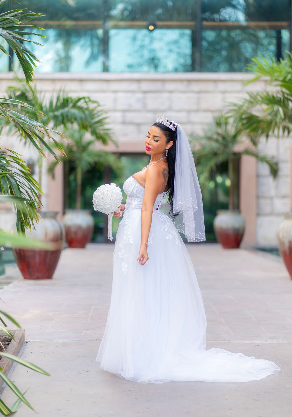 A bride in an elegant white strapless wedding gown and veil poses gracefully among tropical palm plants at an outdoor venue.