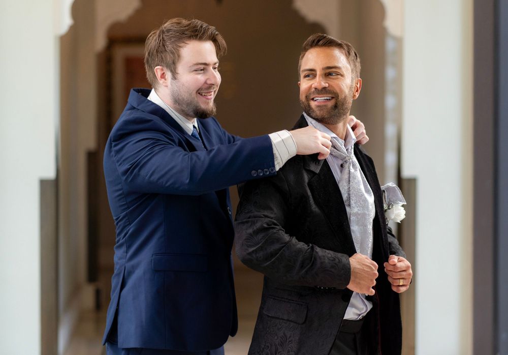 Two men in suits share a lighthearted moment together, with one adjusting the other's tie while both smile warmly.